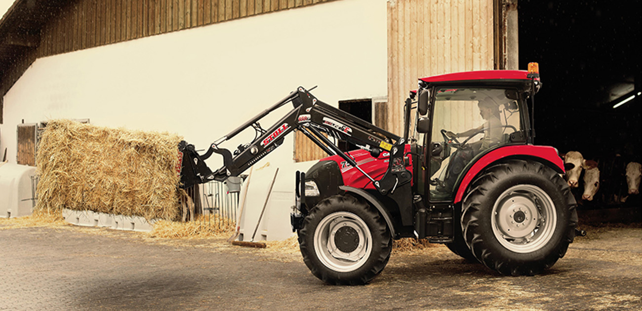 Röd traktor med höbal. I bakgrund lada med kor